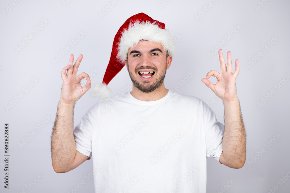 Young handsome man wearing a Santa hat over white background doing ok sign with fingers and smiling, excellent symbol Young handsome man wearing a Santa hat over white background doing ok sign with fingers and smiling, excellent symbol