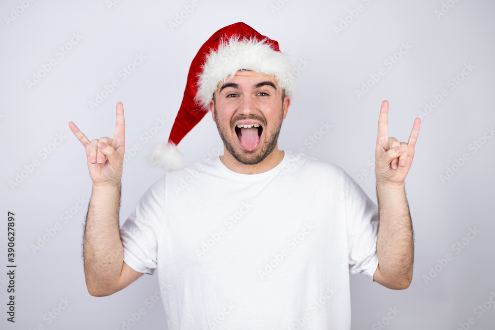 Young handsome man wearing a Santa hat over white background shouting with crazy expression doing rock symbol with hands up Young handsome man wearing a Santa hat over white background shouting with crazy expression doing rock symbol with hands up