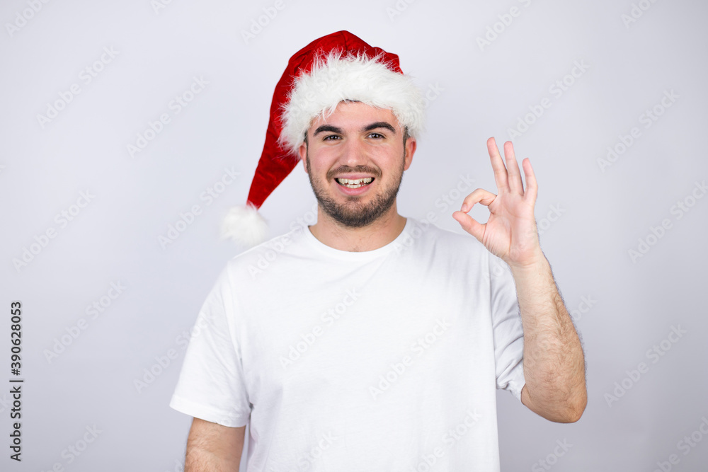 Young handsome man wearing a Santa hat over white background doing ok sign with fingers and smiling, excellent symbol