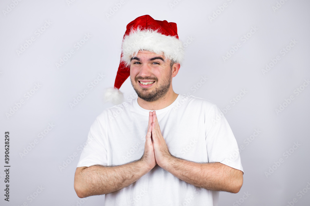 Young handsome man wearing a Santa hat over white background begging and praying with hands together with hope expression on face very emotional and worried Young handsome man wearing a Santa hat over white background begging and praying with hands together with hope expression on face very emotional and worried