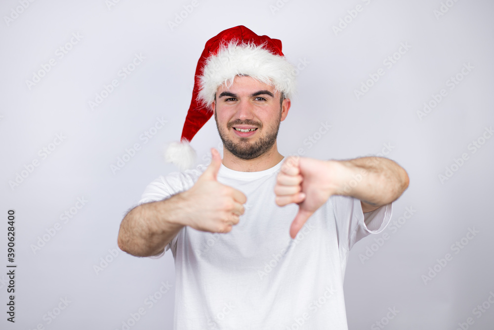 Young handsome man wearing a Santa hat over white background Doing thumbs up and down, disagreement and agreement expression. Crazy conflict Young handsome man wearing a Santa hat over white background Doing thumbs up and down, disagreement and agreement expression. Crazy conflict