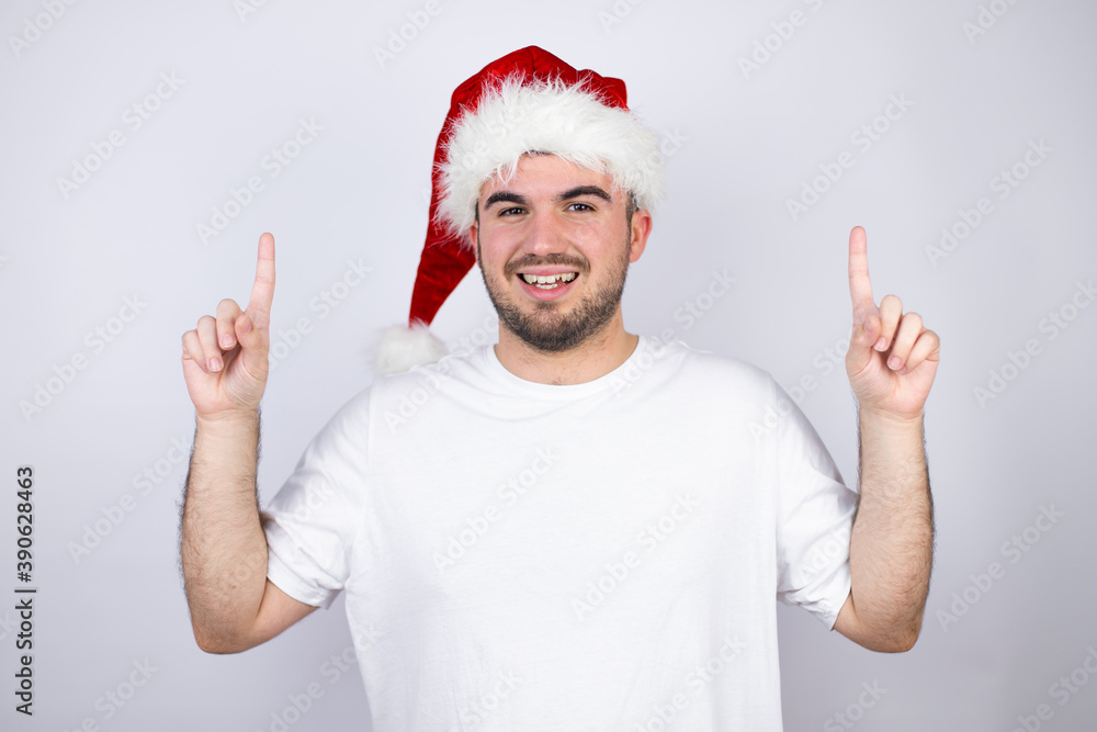 Young handsome man wearing a Santa hat over white background smiling, looking at the camera and pointing up with fingers and raised arms Young handsome man wearing a Santa hat over white background smiling, looking at the camera and pointing up with fingers and raised arms