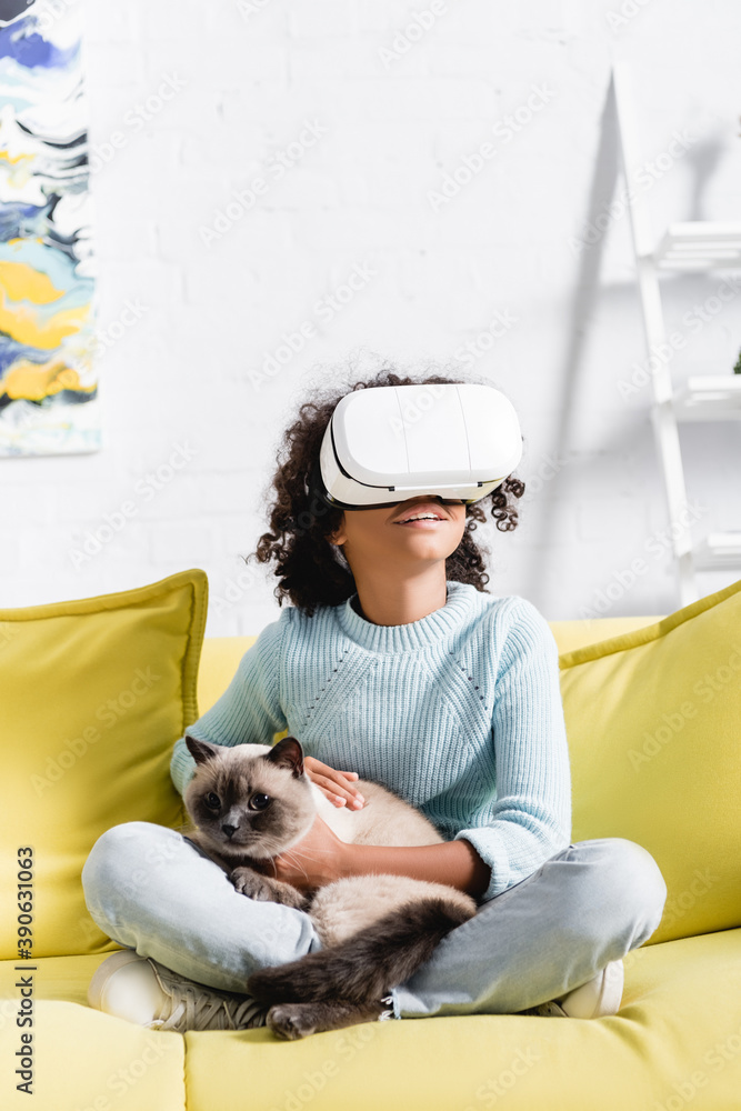 Smiling african american girl with crossed legs wearing vr headset, stroking cat and sitting on sofa on blurred background
