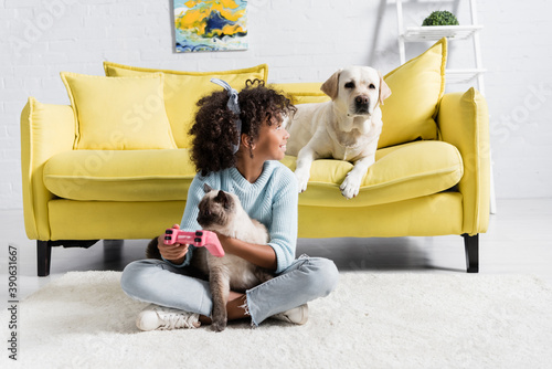 KYIV, UKRAINE - OCTOBER 02, 2020: African american girl with joystick and cat and looking at labrador on sofa at home photo