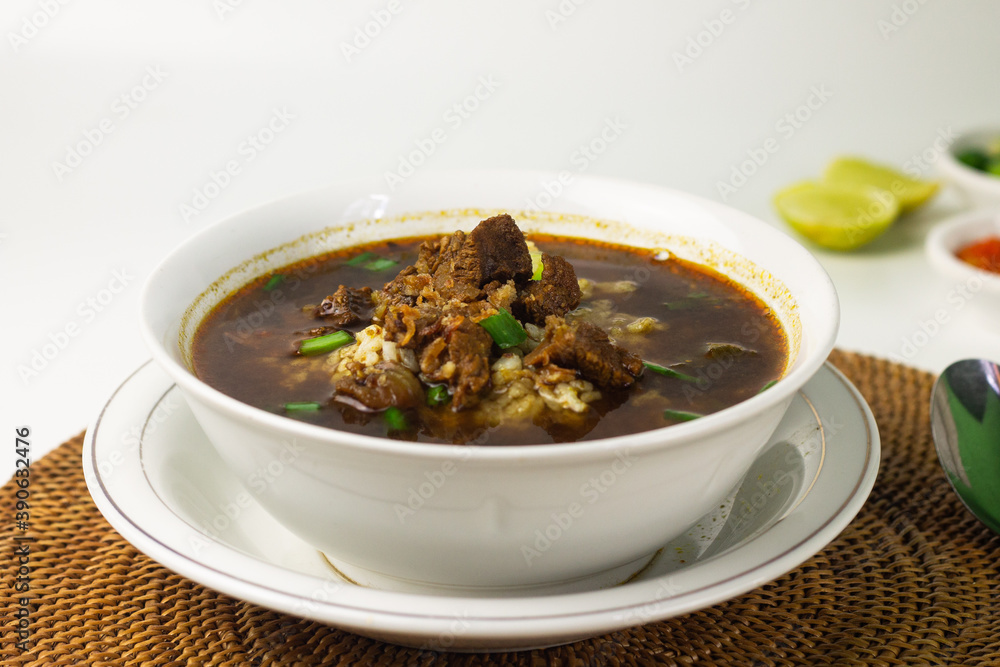 Rawon, Indonesian traditional beef soup from Java Island. Served on white ceramic bowl topped with onion cracker and spring onion,chili paste and lime slices on rattan matt. Isolated white background. Rawon, Indonesian traditional beef soup from Java Island. Served on white ceramic bowl topped with onion cracker and spring onion,chili paste and lime slices on rattan matt. Isolated white background.