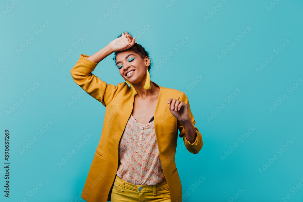 Brunette african girl in yellow jacket smiling with eyes closed. Indoor photo of black young woman dancing on blue background. Brunette african girl in yellow jacket smiling with eyes closed. Indoor photo of black young woman dancing on blue background.