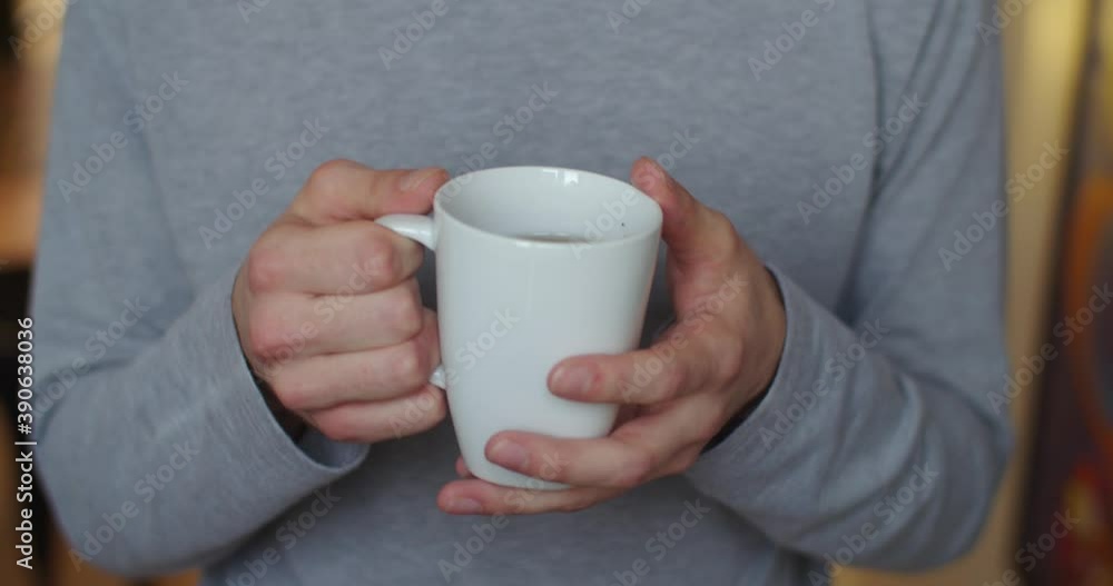 Front view of male hands holding white cup indoors slow motion close up. Faceless man drinking tea enlightened with morning natural light. Breakfast energy healthy lifestyle. Healthcare habits Front view of male hands holding white cup indoors slow motion close up. Faceless man drinking tea enlightened with morning natural light. Breakfast energy healthy lifestyle. Healthcare habits