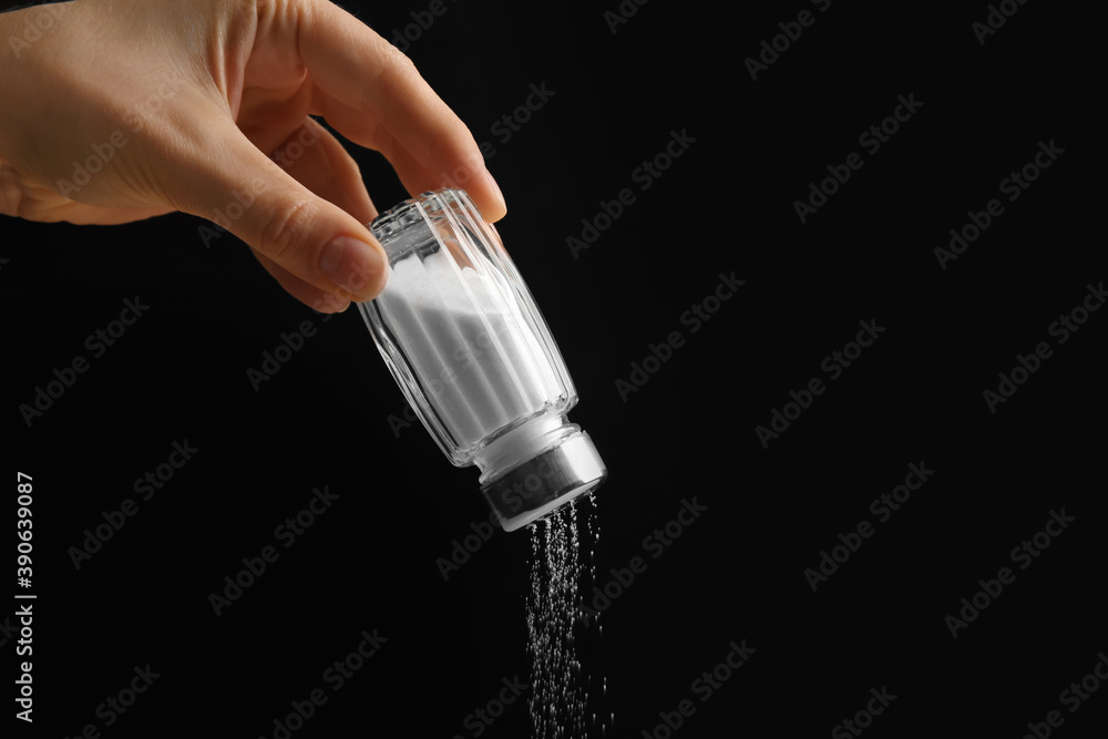 Woman pouring salt from shaker on black background, closeup Woman pouring salt from shaker on black background, closeup