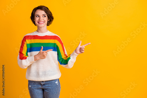 Photo of funky nice brunette hair girl stand point empty space wear rainbow sweater blue jeans isolated on bright yellow background