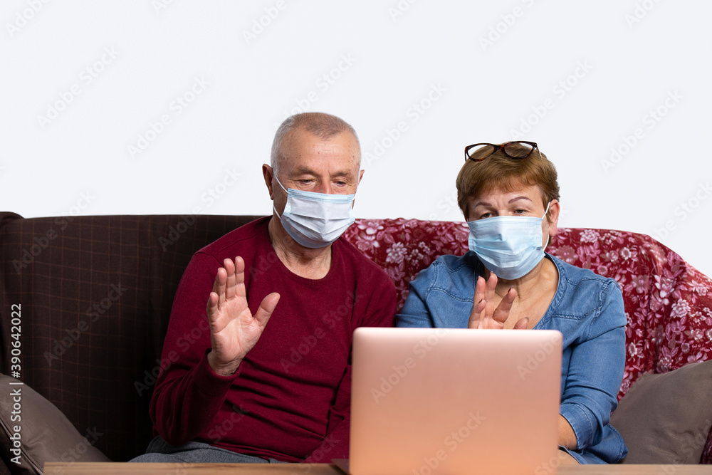 An elderly couple in medical masks waving their hands, looking at the computer screen, talking on a laptop webcam, sitting on the couch at home. Covid 19 and senior online communication concept
