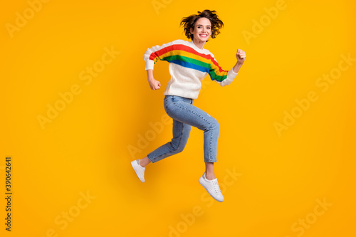 Profile full size photo of cheerful crazy girl jump in air wear rainbow sweater denim jeans white shoes isolated on yellow background