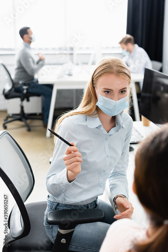 Businesswoman in medical mask holding pen near colleague on blurred foreground