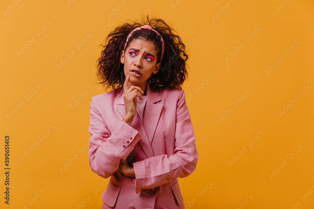 Pensive curly woman in elegant jacket standing on yellow background. Studio photo of worried african lady in pink outfit. Pensive curly woman in elegant jacket standing on yellow background. Studio photo of worried african lady in pink outfit.