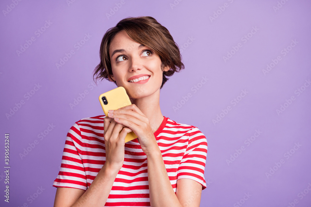 Photo of dreamy funny young girl hold cellphone look up empty space wear striped t-shirt isolated purple color background Photo of dreamy funny young girl hold cellphone look up empty space wear striped t-shirt isolated purple color background