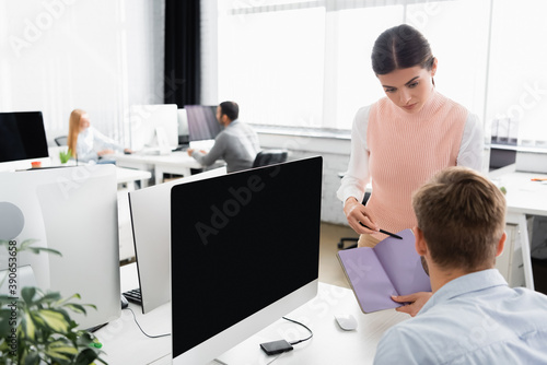 Businesswoman pointing at notebook near colleague and computer with blank screen on blurred foreground