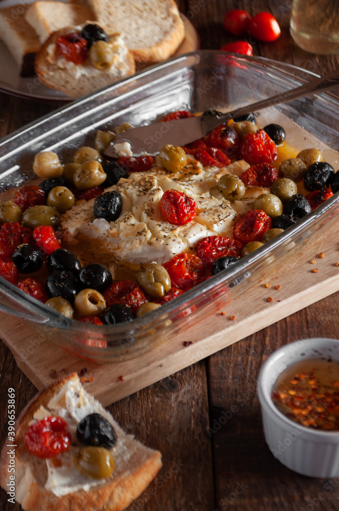 Baked feta cheese with cherry tomatoes, olives, olives, herbs and olive oil, in a glass form, on a wooden background Baked feta cheese with cherry tomatoes, olives, olives, herbs and olive oil, in a glass form, on a wooden background