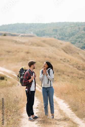 Cheerful african american woman standing near boyfriend with backpack on path during trip