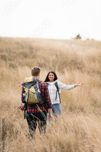 Smiling african american woman smiling at boyfriend with backpack on blurred foreground on grassy hill