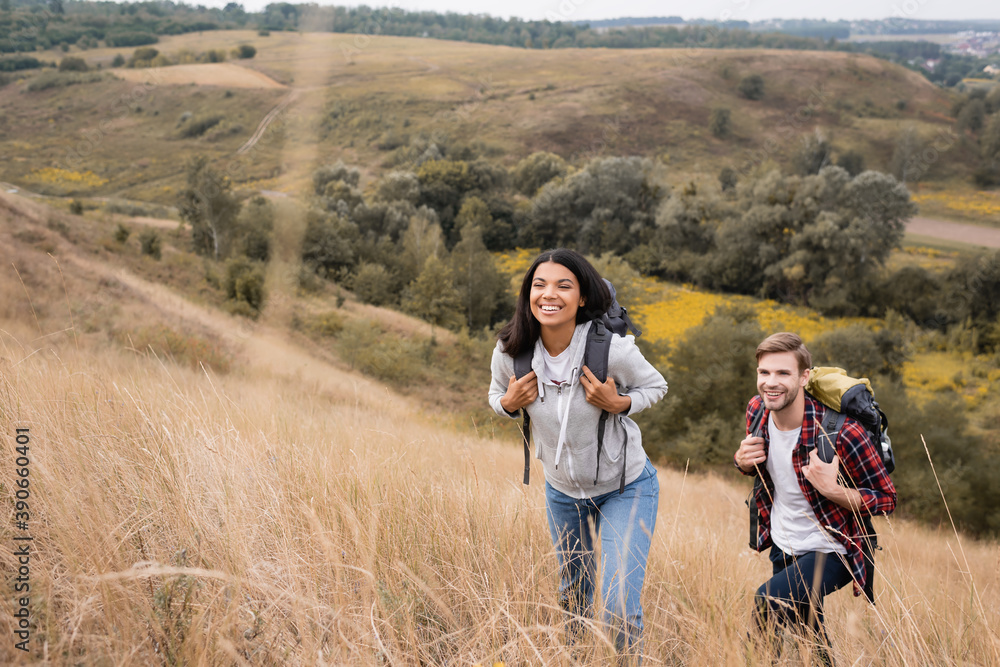 Interracial couple with backpacks walking on hill with grass during trip