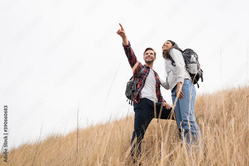 Low angle view of smiling man with backpack pointing with finger away near african american woman while standing on grassy hill with sky at background