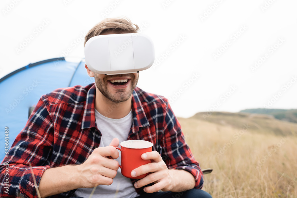 Smiling man in vr headset holding cup near tent on blurred background on meadow Smiling man in vr headset holding cup near tent on blurred background on meadow