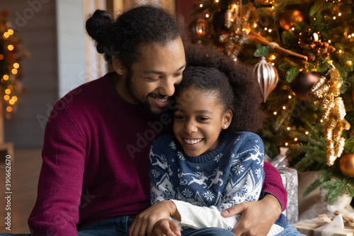 Close up happy African American man and adorable daughter hugging, celebrating Christmas, sitting near festive tree at home, smiling little girl and dad wearing sweaters cuddling, having fun