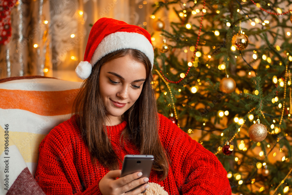 Smiling girl in red hat and sweater scrolling social media in her mobile phone. Cheerful girl sitting in comfortable armchair against glimmering Christmas tree and surfs the Internet Smiling girl in red hat and sweater scrolling social media in her mobile phone. Cheerful girl sitting in comfortable armchair against glimmering Christmas tree and surfs the Internet