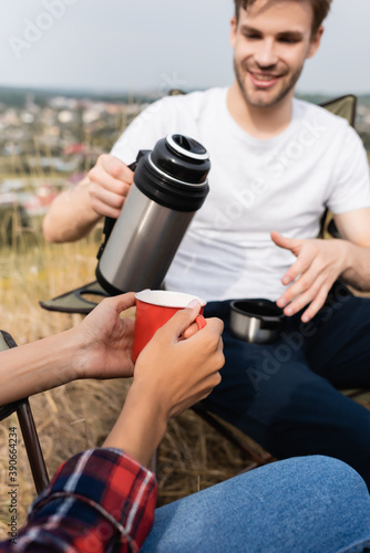 African american woman holding cup near smiling man with thermos on blurred foreground during camping