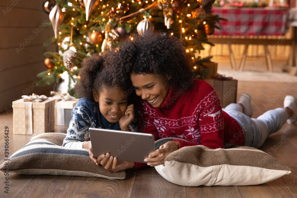 Smiling African American woman with daughter using tablet, having fun with device, lying on pillows on warm floor near Christmas tree at home, happy family shopping online, choosing gifts
