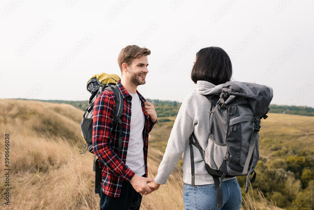 Smiling man holding hand of african american girlfriend with backpack on grassy lawn Smiling man holding hand of african american girlfriend with backpack on grassy lawn