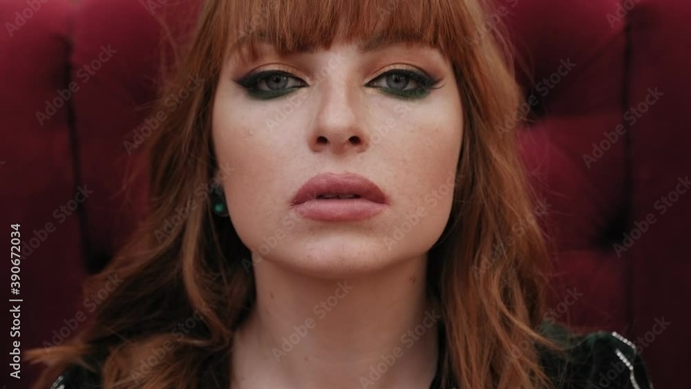 Close up of the face of a red-haired girl with freckles on her face, she sits in a red soft chair and looks at the camera. Close up of the face of a red-haired girl with freckles on her face, she sits in a red soft chair and looks at the camera.