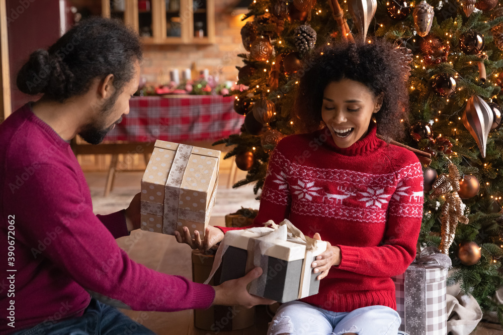 Overjoyed young African American man and woman giving Christmas gift boxes, presents, sitting on floor near decorated festive tree, family celebrating New Year at home, enjoying winter holidays - obrazy, fototapety, plakaty 