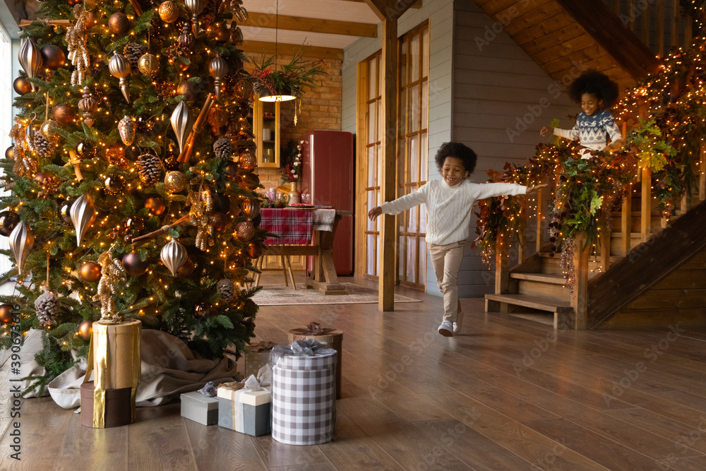 Overjoyed African American little kids wearing sweaters running downstairs on Christmas morning to opening presents, gift boxes under decorated festive tree, excited girl and boy celebrating New Year - obrazy, fototapety, plakaty 