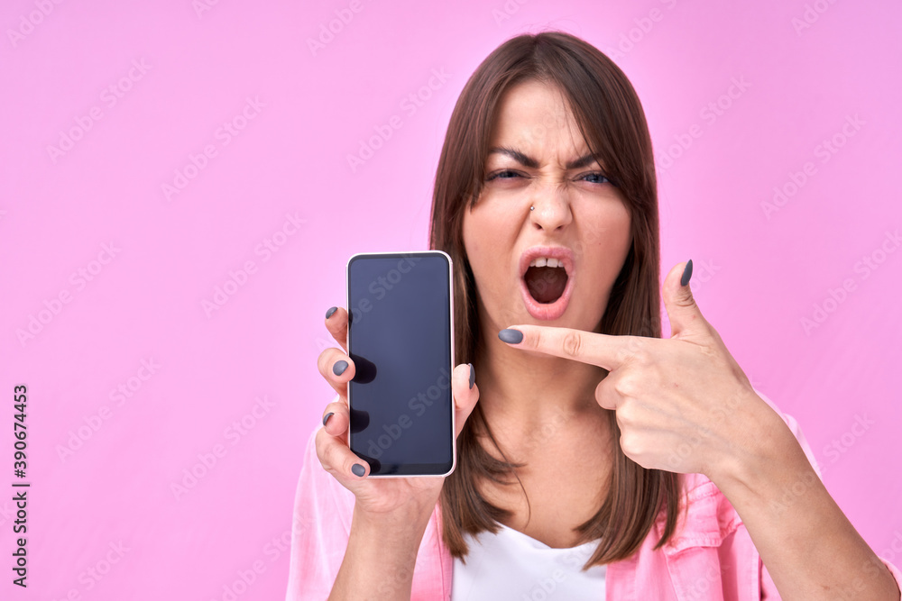 Brunette happy girl with amazed open mouth points finger at blank mobile phone screen with copy space isolated in pink studio Brunette happy girl with amazed open mouth points finger at blank mobile phone screen with copy space isolated in pink studio