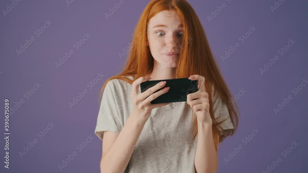 A happy young woman is playing games on her smartphone standing isolated over a violet background A happy young woman is playing games on her smartphone standing isolated over a violet background