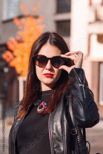 Beautiful woman in a black leather jacket with red lips. A woman in sunglasses corrects them with her hand. Beautiful woman on a background of autumn city