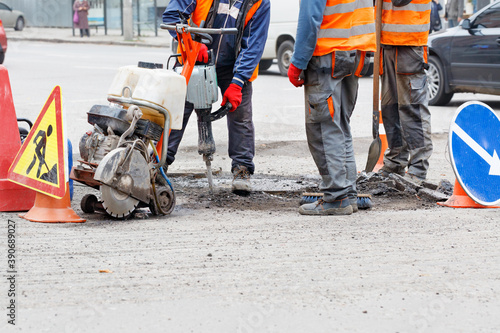A team of road workers repairs a section of road with an electric jackhammer, petrol cutter, shovel and brush.