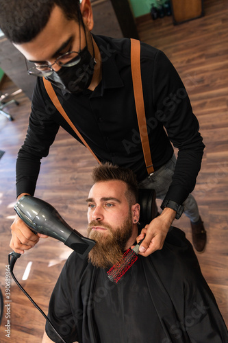 A bearded boy has his beard combed by a barber with a comb and a hairdryer, and mask for pandemic protection. photo