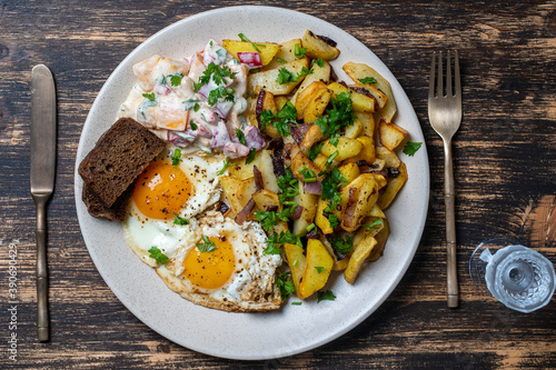 Ukrainian food, fried potatoes with onions, eggs, vegetable salad, black bread and glass of vodka on wooden background