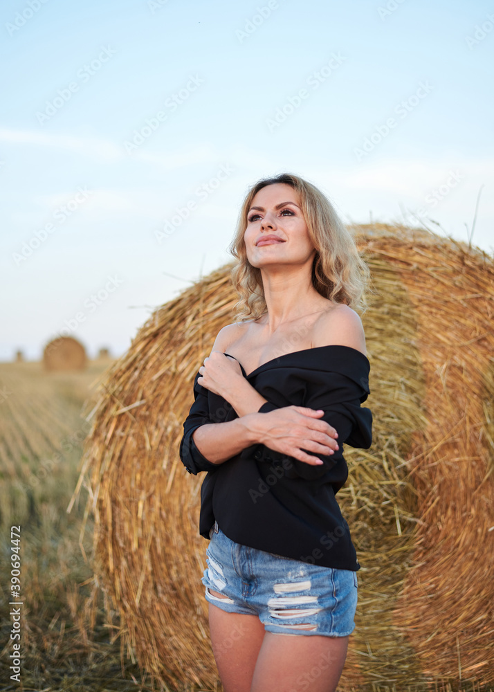 Young blond woman, wearing black jacket and jeans shorts, standing by wheat bale on rural field on summer evening. Three-quarter female portrait during sunset at natural landscape. Young blond woman, wearing black jacket and jeans shorts, standing by wheat bale on rural field on summer evening. Three-quarter female portrait during sunset at natural landscape.