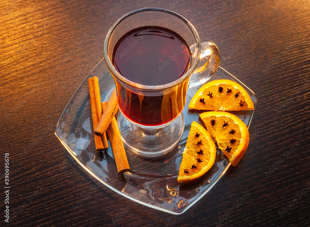 Tea in a glass cup on a saucer next to orange slices with cloves and cinnamon sticks. Wooden table as a background