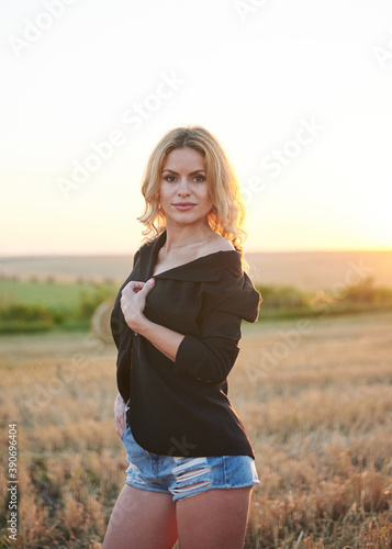 Young blond woman, wearing black jacket and jeans shorts, posing on wheat field on summer evening. Creative stylish three-quarter female portrait during sunset at natural rural landscape. © Natalia