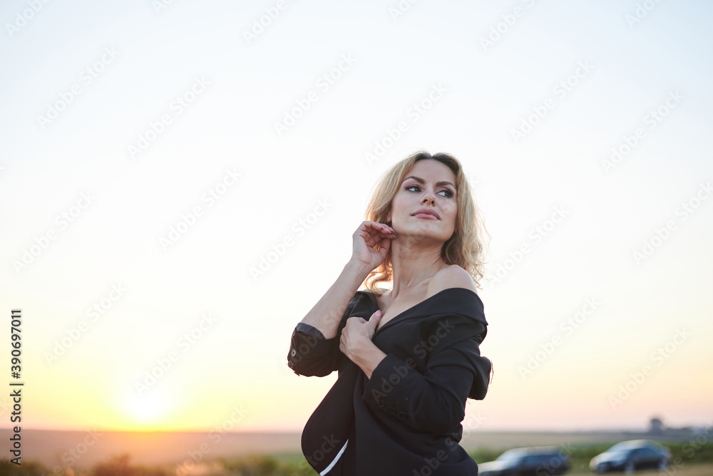 Young blond woman, wearing black jacket and jeans shorts, posing on wheat field on summer evening. Creative stylish three-quarter female portrait during sunset at natural rural landscape.