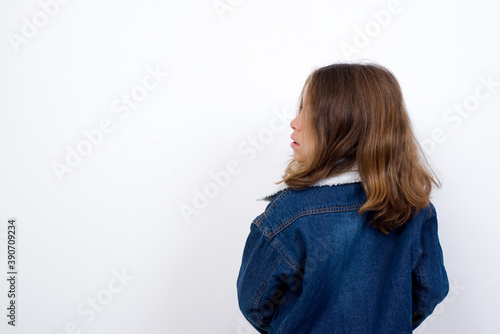 The back side view of a Little caucasian girl with beautiful blue eyes wearing denim jacket standing over isolated white background . Studio Shoot.