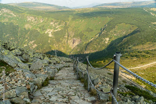 A steep mountain trail with handrails on chains, bright mountains in the background.