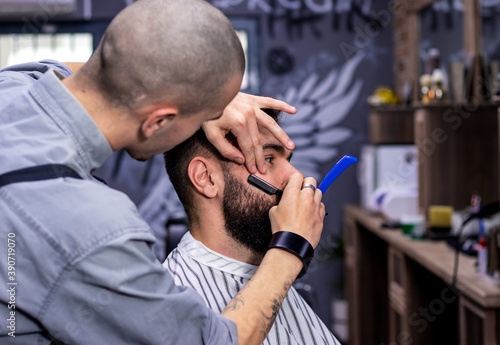 Young bearded man getting haircut by professional at modern barbershop. Beard and sideburns shaving and shaping with vintage straight razor. Barber shop advertising concept. Movember.