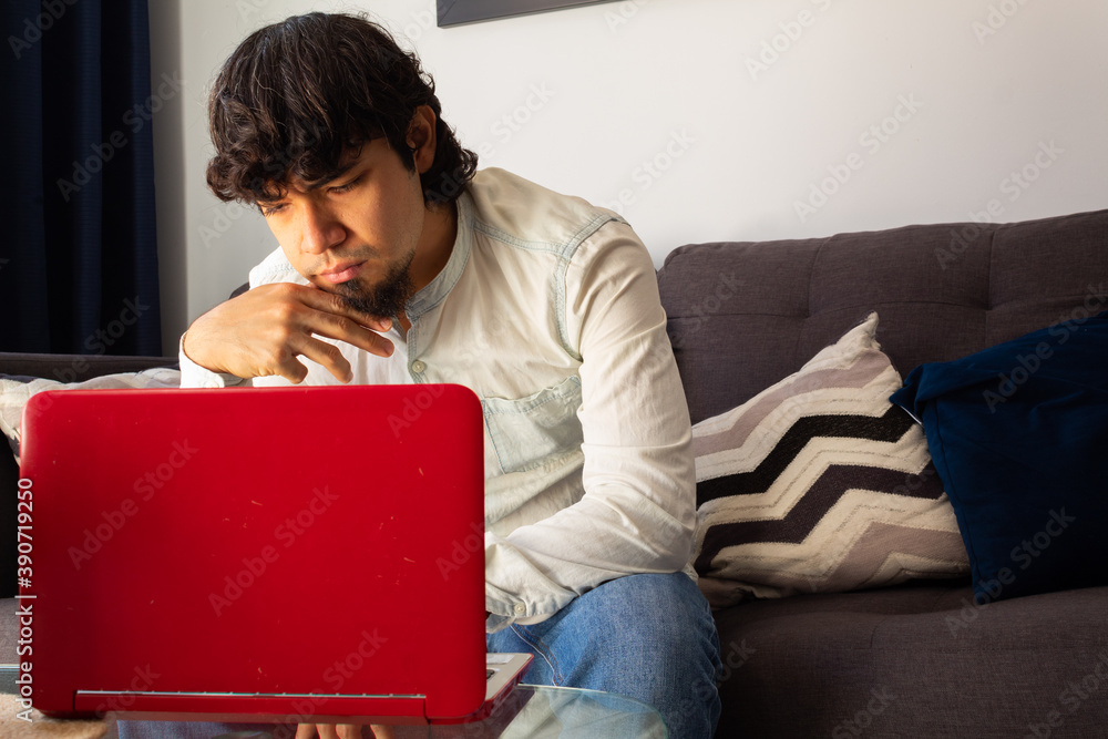 Hispanic young man in casual clothes brainstorming at a distance with a laptop on a sofa with a calm and thoughtful attitude