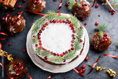 Small chocolate muffins and big cake are poured with chocolate, decorated with winter berries. Cinnamon sticks, snowflake decor on a dark table.