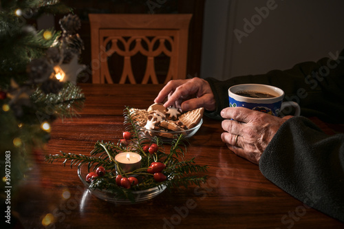 Hands of an elderly single man sitting alone at a table with Christmas cookies  coffee and festive decoration next to an empty chair  lonely holidays during the croronavirus pandemic or after a loss