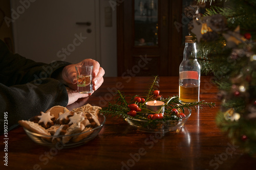 Hands of an elderly man holding a shot glass alone at a table with Christmas decoration  cookies  candle and a half-empty bottle of liquor  lonely holidays during the covid-19 pandemic  copy space